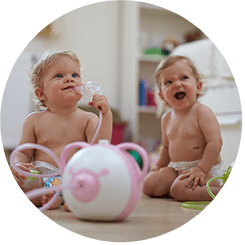 Due bambine felici siedono sul pavimento con l'aspiratore nasale elettrico Nosiboo Pro