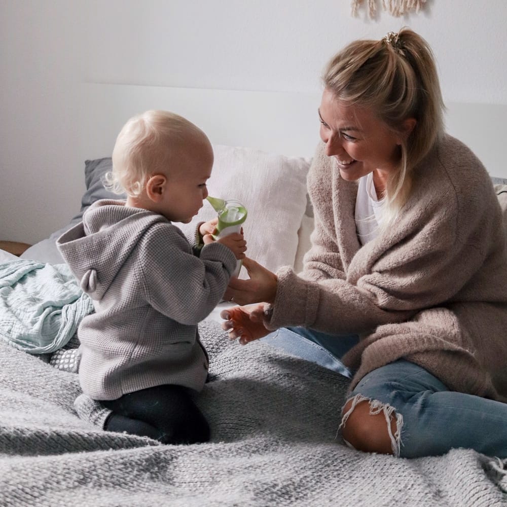 Mamma e suo figlio seduti su un letto e aspirano il naso del bambino con un aspiratore nasale elettrico e portatile Nosiboo Go