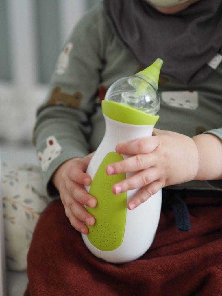 Un bambino carino siede sul letto e tiene l'aspiratore nasale elettrico Nosiboo Go in mano