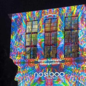 Il logo di Nosiboo visualizzato con luci colorate su un edificio, per informare del livello di sponsorizzazione di bronzo dello Zsolnay Light Festival di Pécs.