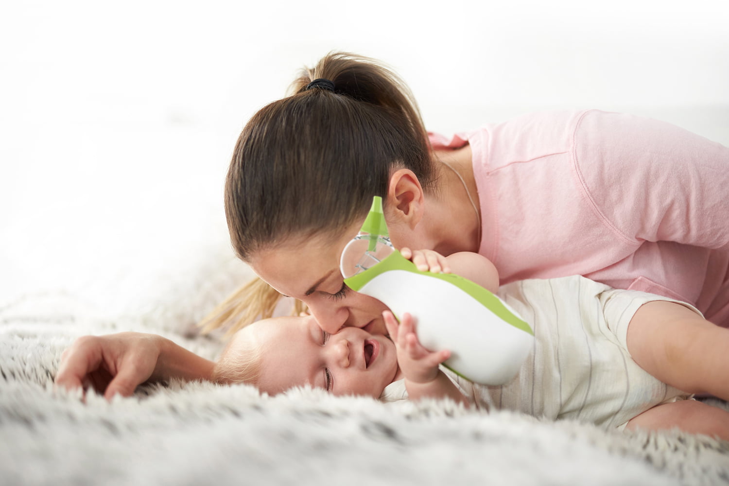 A mom kissing her baby boy who is carrying the Nosiboo Go portable nasal aspirator in his hands while lying on the bed