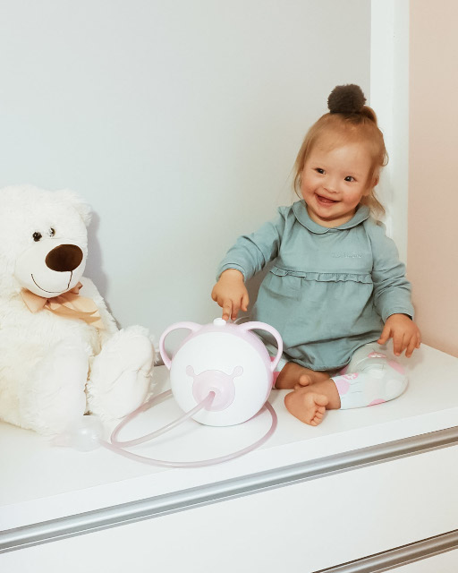Una bambina sorridente con un aspiratore nasale elettrico Nosiboo Pro in colore rosa e un orsetto di peluche