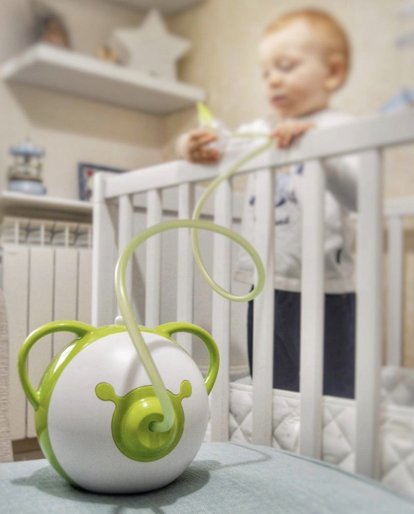 Aspiratore nasale elettrico Nosiboo Pro verde, con un bambino in piedi su un letto sullo sfondo